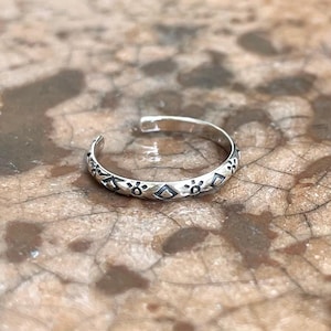 Può includere: Un anello per l'alluce color argento con un design aperto. L'anello presenta un motivo ripetuto di forme floreali e a diamante attorno alla fascia. L'anello è esposto su una superficie marrone strutturata.