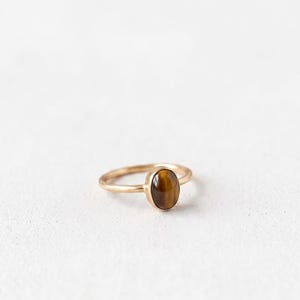 Könnte beinhalten: Ein goldener Ring mit einem ovalen Tigerauge-Stein. Der Ring hat ein dünnes Band und eine Lünettenfassung für den Edelstein. Der Tigerauge-Stein hat eine warme braune Farbe mit Chatoyanz. Der Ring ist vor einem weißen Hintergrund platziert.