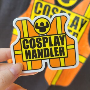 May include: A white sticker with a yellow and orange design. The sticker features a cartoon figure with raised arms and the text "Cosplay Handler".