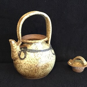May include: A small, speckled, beige teapot with a brown handle and a brown lid. The teapot has a brown and black design around the middle.