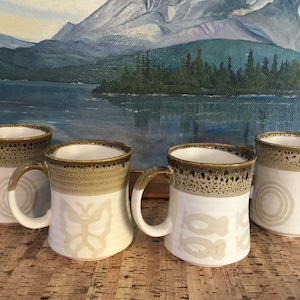 May include: Set of four ceramic mugs with brown speckled glaze and white designs. Each mug features a different design: a butterfly, circles, and two abstract shapes.