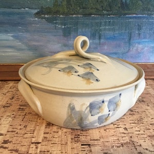 May include: A white ceramic casserole dish with a lid. The dish has two handles and a unique swirl handle on the lid. The dish is decorated with a blue and yellow fish design.
