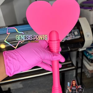 Puede incluir: Un abanico rosa brillante en forma de corazón con un mango largo. El abanico lo sostiene una persona con guantes rosas. El texto "GENESIS.PRINTS & CUSTOMS" es visible en la imagen.
