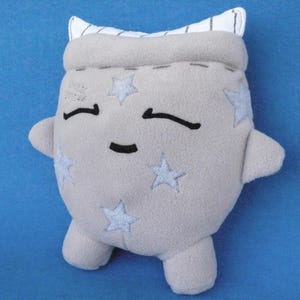Puede incluir: Un peluche con forma de figura redondeada y cuerpo de forro polar gris claro, con ojos y una pequeña boca bordados en negro. La superficie está decorada con aplicaciones de estrellas azul claro y la parte superior tiene un borde de tela blanca.