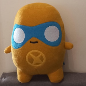 May include: A mustard yellow plush toy shaped like a rounded character with small arms and legs. It features a blue mask across the eyes with two white circular eye patches, a small black stitched mouth, and a circular emblem on the chest.