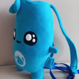 Puede incluir: Una mochila de peluche azul brillante con forma de personaje, con grandes ojos bordados en blanco y negro, una pequeña boca cosida y un logotipo circular blanco en el frente. Presenta una construcción de tela suave, asa y correas azules.