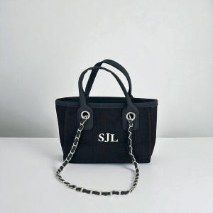 Puede incluir: Bolso tote negro con una correa de cadena y asas negras. El bolso presenta las iniciales "SJL" en letras blancas en la parte delantera. El bolso tiene forma rectangular y una correa de cadena plateada.