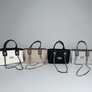 May include: Four canvas tote bags with chain straps and top handles. Each bag has a different colour scheme: white with black trim, tan with brown trim, black with black trim, and grey with grey trim. Each bag has initials printed on the front.