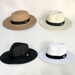 Op de afbeelding: Vier fedora hoeden in beige, crème, zwart en wit, elk voorzien van een zwarte band met goud- of witgeborduurde initialen. De hoeden hebben een gevlochten textuur, een ingedeukte kroon en een brede rand, met de letters SI, AMS, ZY en HP.