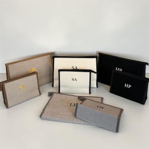 Könnte beinhalten: Eine Sammlung personalisierter Kosmetiktaschen in verschiedenen Größen und Farben. Die Taschen sind in Beige, Schwarz und Grau gehalten, mit kontrastierenden Zierleisten und gold- oder weißgestickten Initialen. Die Taschen sind auf einer weißen Oberfläche angeordnet.