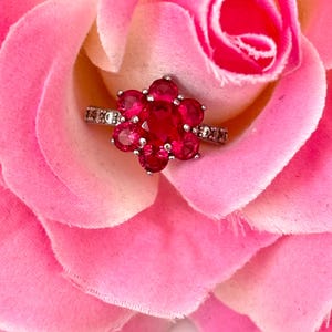 Op de afbeelding: Een zilveren ring met een bloemvormig ontwerp, bezet met rode edelstenen. De ring is geplaatst op een roze roos, wat een romantische scène creëert. De band van de ring is versierd met kleine, heldere stenen. Close-up.