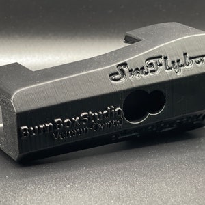 May include: A black, 3D-printed object with the text "SmFlyboy" and "Burn Box Studio" visible. The object has a rectangular shape with a curved top and a hole in the side. The object appears to be a custom-made part.