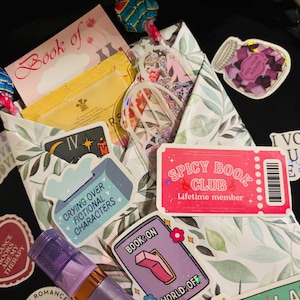 May include: A gift package wrapped in white paper with green leaf patterns, decorated with various stickers including a pink Spicy Book Club ticket, a purple Book On World Off sticker, and a heart-shaped Romance Books Are My Therapy sticker.