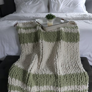 May include: A white and sage green chunky knit throw blanket with a textured, cable knit pattern. The blanket is draped over a white bed with white pillows and a small potted plant on a silver tray.