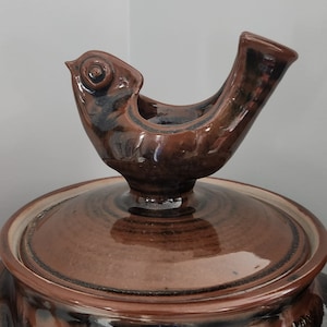 Könnte beinhalten: Eine braune Keramikdose mit einem Deckel in Form eines Vogels. Die Dose hat eine dunkelbraune Glasur mit einem helleren braunen Streifen um die Mitte. Der Vogel hat eine schwarze und braune Glasur und sitzt auf dem Deckel.