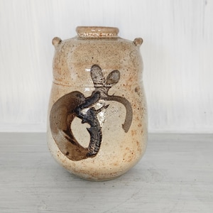 May include: A small, brown ceramic jar with a unique design. The jar has a rounded shape and two small handles on the sides. The design features a dark brown swirl pattern.