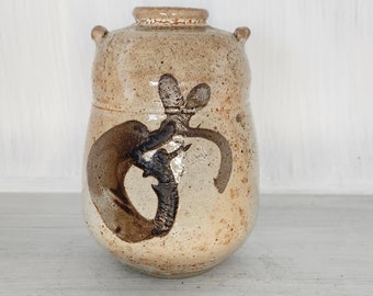 May include: A small, brown ceramic jar with a unique design. The jar has a rounded shape and two small handles on the sides. The design features a dark brown swirl pattern.