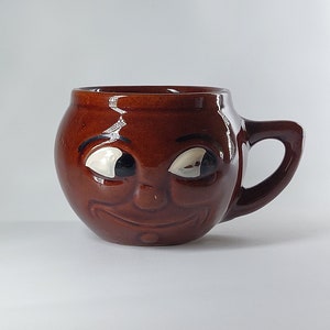 May include: A brown ceramic mug shaped like a face with white eyes and a smile. The mug has a handle on the right side.