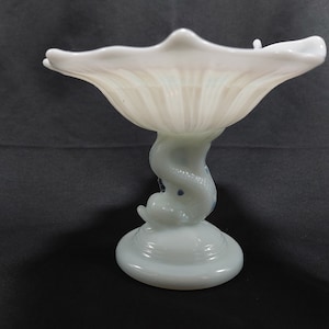 Puede incluir: Un cuenco de vidrio blanco con un borde festoneado se asienta sobre una base de vidrio blanco con forma de sirena. La base tiene lunares azules y blancos.