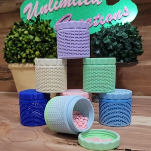 May include: A collection of colorful, small, lidded containers in shades of blue, green, purple, and cream. One container is open, revealing pink heart-shaped candies. The containers have a textured, woven-like design.