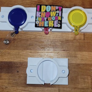 May include: A white badge holder with a blue and yellow retractable badge reel. The badge holder has the text "I DON'T KNOW I JUST WORK HERE" on a black background. The badge holder is on a wooden surface.