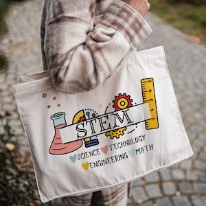 May include: A white canvas tote bag with a colorful STEM design. The design includes a beaker, lightbulb, gears, and a ruler. The words "STEM", "Science", "Technology", "Engineering", and "Math" are printed on the bag.