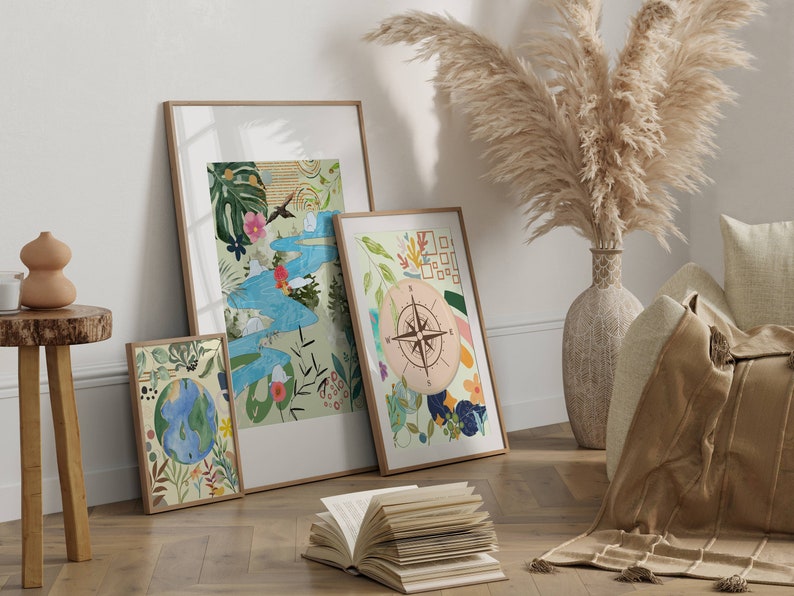 May include: Three framed art prints with a nature theme. The prints feature a compass, a globe, and a stylized landscape with a river and birds. The prints are displayed on a white wall with a vase of dried pampas grass in the background.