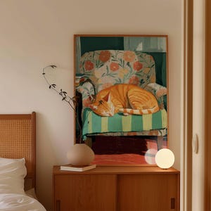 Può includere: Una stampa artistica incorniciata raffigura un gatto rosso addormentato su una poltrona floreale. L'opera d'arte presenta una calda tavolozza di colori e una cornice in legno. Un mobile in legno con un vaso e una lampada è in primo piano.