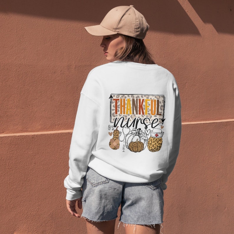 May include: White sweatshirt with a graphic print of a stethoscope, a pumpkin, and a bandage with the text "Thankful Nurse" in a leopard print pattern.