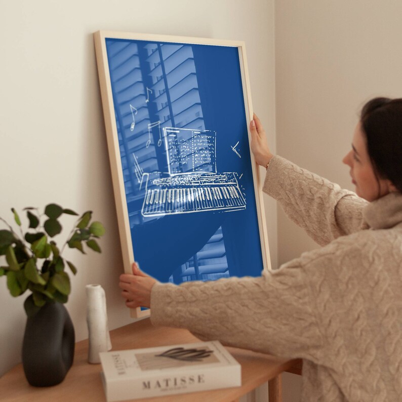 May include: A person hangs a framed print on a wall. The print is on a blue background with a white outline of a keyboard and musical notes.