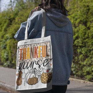 May include: A white tote bag with the text "THANKFUL nurse" in orange and yellow letters. The bag has a leopard print design and features a small image of a nurse's hat with a red cross and a pumpkin with a stethoscope.