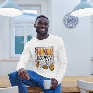 May include: White long-sleeved shirt with a graphic print of a leopard print background with the text "Thankful Nurse" in orange and brown letters. The graphic also includes a pumpkin, a gourd, and a stethoscope with a leopard print design.