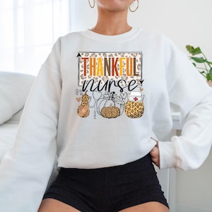 May include: White sweatshirt with the text "Thankful Nurse" in a leopard print frame. The design includes a stethoscope, a pumpkin, and a gourd with a bandage.