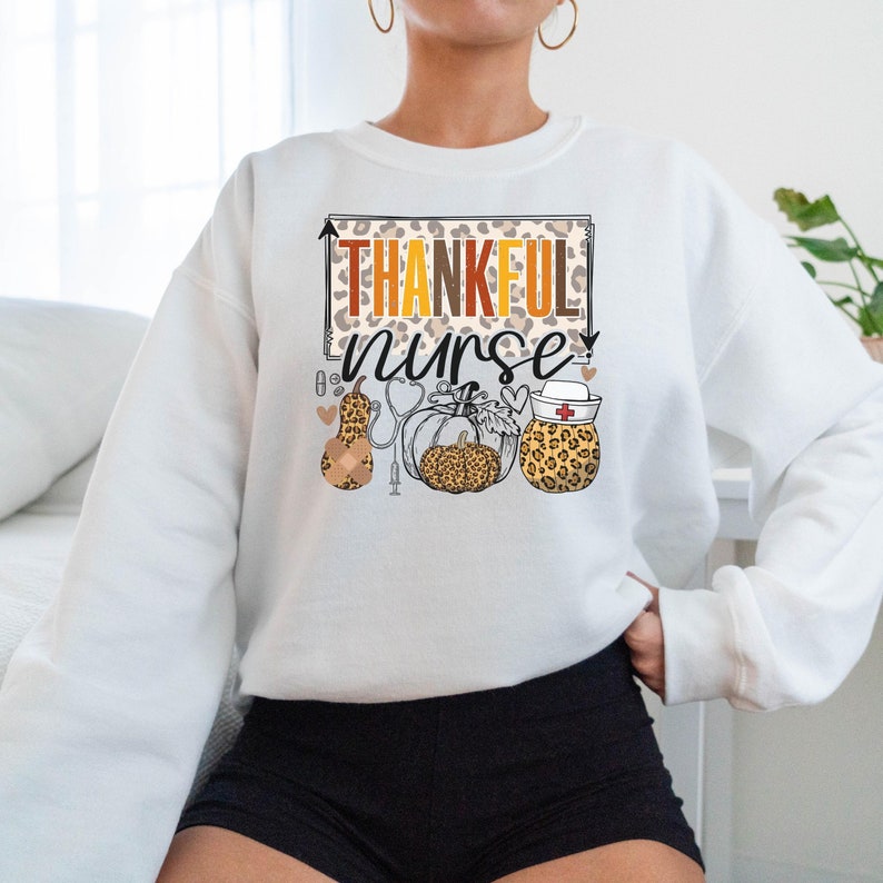 May include: White sweatshirt with the text "Thankful Nurse" in a leopard print frame. The design includes a stethoscope, a pumpkin, and a gourd with a bandage.