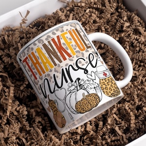 May include: White ceramic coffee mug with a brown, orange, and yellow "Thankful Nurse" design. The design features a stethoscope, a syringe, a heart, and a pumpkin with a leopard print.