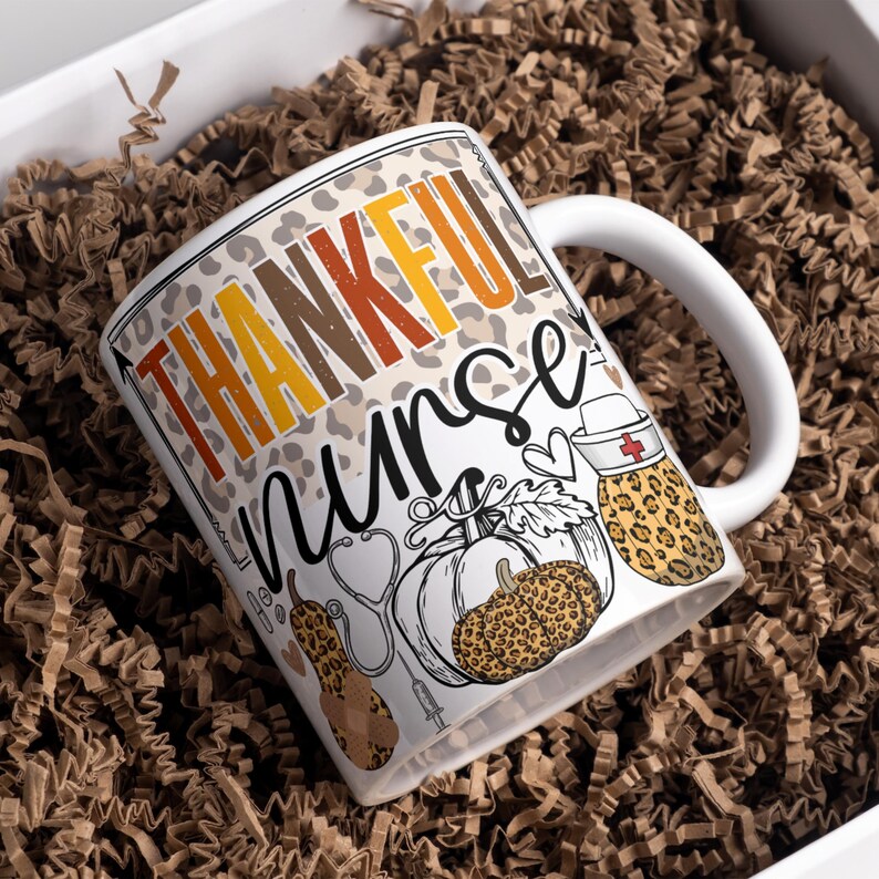 May include: White ceramic coffee mug with a brown, orange, and yellow "Thankful Nurse" design. The design features a stethoscope, a syringe, a heart, and a pumpkin with a leopard print.