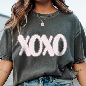 Puede incluir: Camiseta gris oscuro con la palabra "XOXO" en un dise&ntilde;o a cuadros rosa con un borde de encaje blanco. Una prenda informal, adecuada para el uso diario.