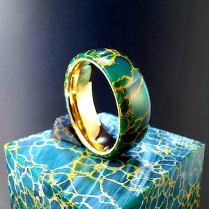 Puede incluir: Un anillo verde y dorado con un patrón de mármol. El anillo está hecho de carburo de tungsteno y está pulido a alto brillo.