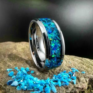 Könnte beinhalten: Ein silberner Ring mit einer blauen und grünen Opal-Einlage. Der Ring liegt auf einem Bett aus kleinen blauen und grünen Opal-Stücken.