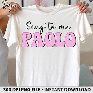 May include: A white short-sleeved t-shirt featuring the dark green text Sing to me and the large pink text PAOLO in a rounded font. The shirt has a subtle white hexagonal pattern printed across the fabric and a crew neckline.