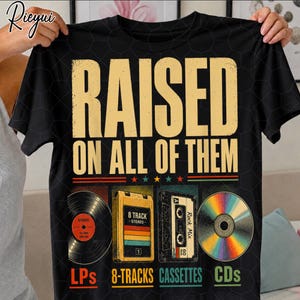 May include: A black short-sleeve t-shirt featuring the text RAISED ON ALL OF THEM in large yellow letters. Below the text are illustrations of a vinyl record, an 8-track tape, a cassette tape, and a CD, with labels LPs, 8-TRACKS, CASSETTES, and CDs.