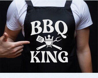 Può includere: Grembiule nero con la scritta bianca "BBQ KING" e un disegno di griglia, spatola e forchetta. Una persona con una maglietta bianca indica il grembiule.