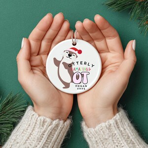 Könnte beinhalten: Weißer Keramik-Weihnachtsschmuck mit einem Cartoon-Otter mit Weihnachtsmütze. Der Schmuck hat den Text "Otterly Amazing OT" mit dem Namen "Megan" und dem Jahr "2025". Der Schmuck wird in zwei Händen gehalten.
