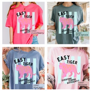 K&ouml;nnte beinhalten: Eine Kollektion von vier kurz&auml;rmeligen T-Shirts in den Farben Crunchberry, Blue Spruce, Blue Jean und Blossom. Jedes Shirt zeigt eine Grafik eines rosa Tigers mit dem Text EASY TIGER und EVERYONE LEARNS DIFFERENTLY auf hellblauen Streifen.