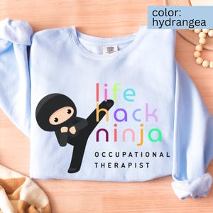 Może przedstawiać: Błękitna bluza z grafiką ninja. Projekt przedstawia kreskówkowego ninja kopiącego, ze słowami "life hack ninja" w kolorowych literach i "Occupational Therapist" poniżej.