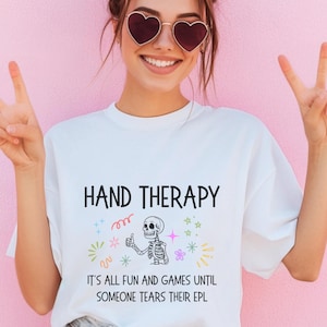 May include: White t-shirt with the words "HAND THERAPY" in black. Below the text is a skeleton giving a thumbs up, surrounded by colourful graphics. The text "IT'S ALL FUN AND GAMES UNTIL SOMEONE TEARS THEIR EPL" is printed below the skeleton.