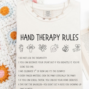 May include: A cream-coloured t-shirt with the text "HAND THERAPY RULES" in black. The shirt features illustrations and humorous rules for hand therapy, including "DO NOT LICK THE THERAPUTTY." A silver butterfly necklace is draped on the shirt.