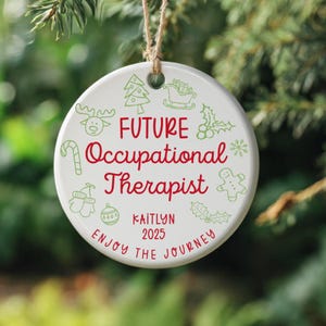 Könnte beinhalten: Ein weißer Keramik-Weihnachtsschmuck mit rotem und grünem Design. Der Schmuck trägt die Aufschrift "FUTURE Occupational Therapist" mit dem Namen "Kaitlyn" und dem Jahr "2025". Der Schmuck hat eine Schnur zum Aufhängen und ist von grünem Laub umgeben.
