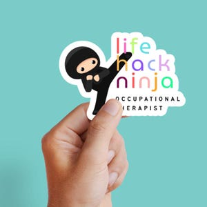 May include: A sticker showing a cartoon ninja in a black outfit, kicking. The sticker has the words "life hack ninja" in colourful letters, with "Occupational Therapist" below. The sticker is held in a hand against a teal background.