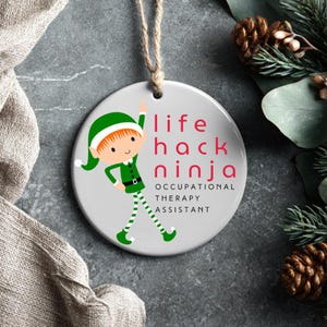 Może przedstawiać: Okrągła, ceramiczna ozdoba świąteczna z postacią elfa z kreskówki. Elf ma na sobie zielony kapelusz, kurtkę i buty oraz zielono-białe spodnie w paski. Tekst brzmi "life hack ninja OCCUPATIONAL THERAPY ASSISTANT."